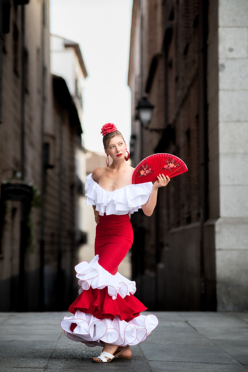 A dancer in a traditional red and white flamenco dress twirls gracefully while holding a red fan in a narrow European alley.