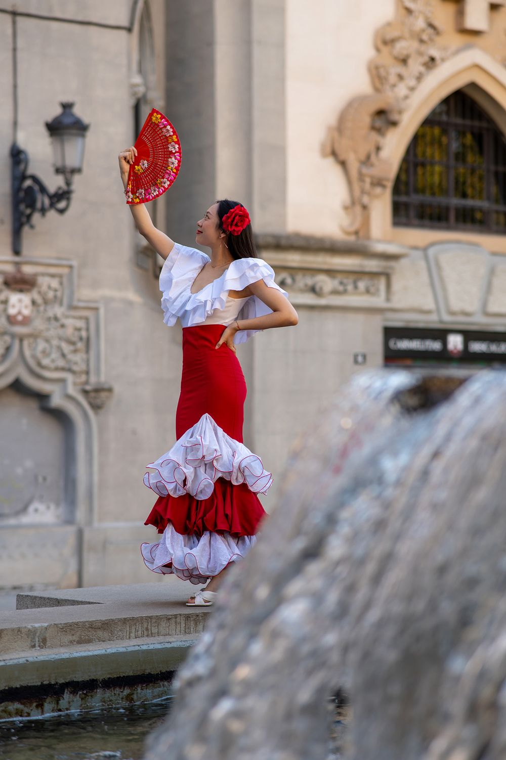 Flamenco dancer in traditional red and white ruffled dress poses gracefully by fountain in ornate European courtyard.