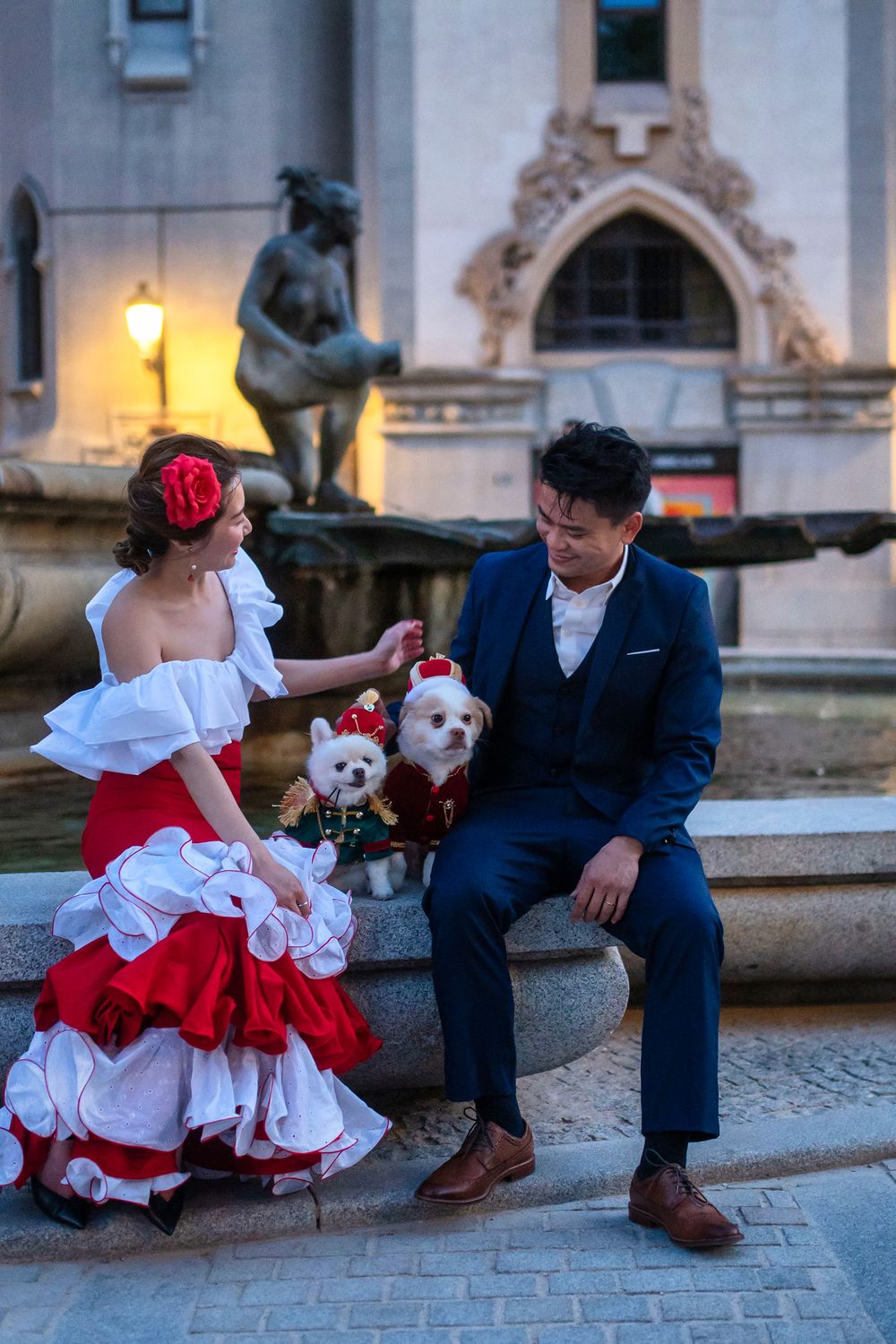 A couple in formal attire sits on stone steps with two small white dogs dressed in festive outfits by a statue.