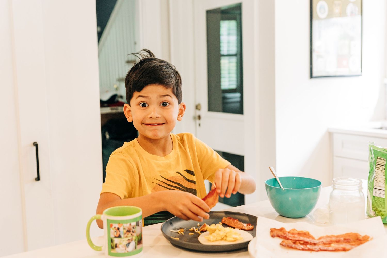 Smiling youngster in yellow shirt sits at kitchen table with breakfast plate and turquoise bowl nearby.