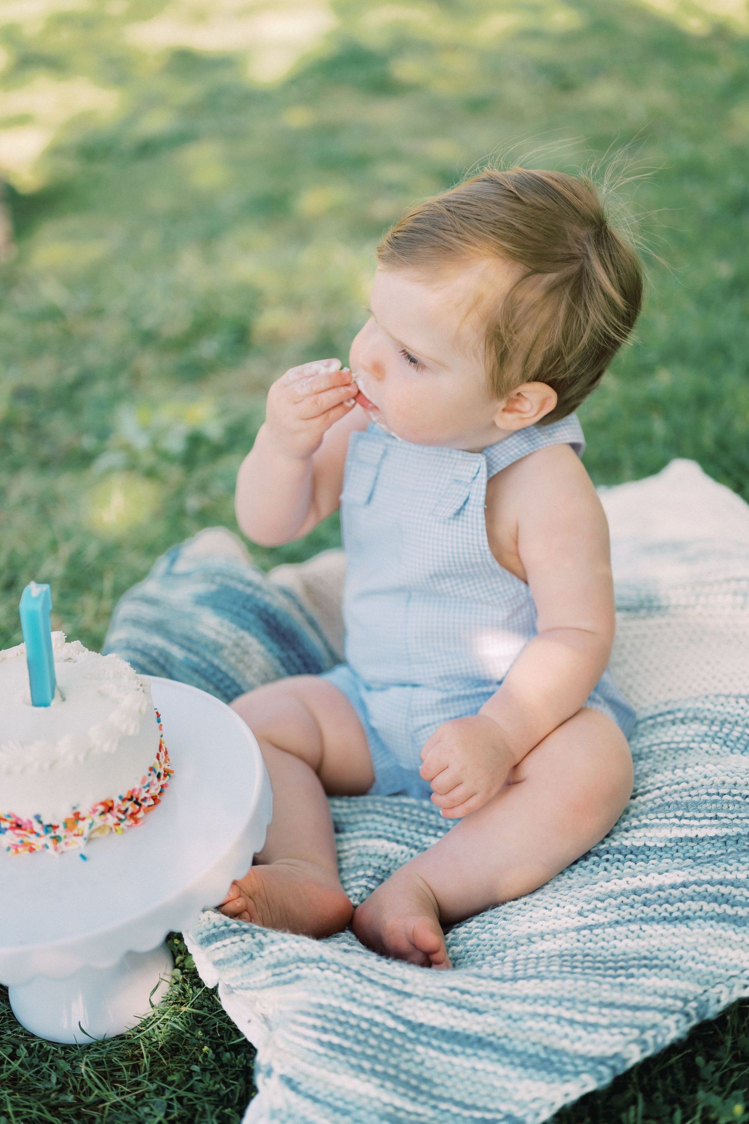 Baby in blue romper sits near decorated birthday cake on striped blanket.
