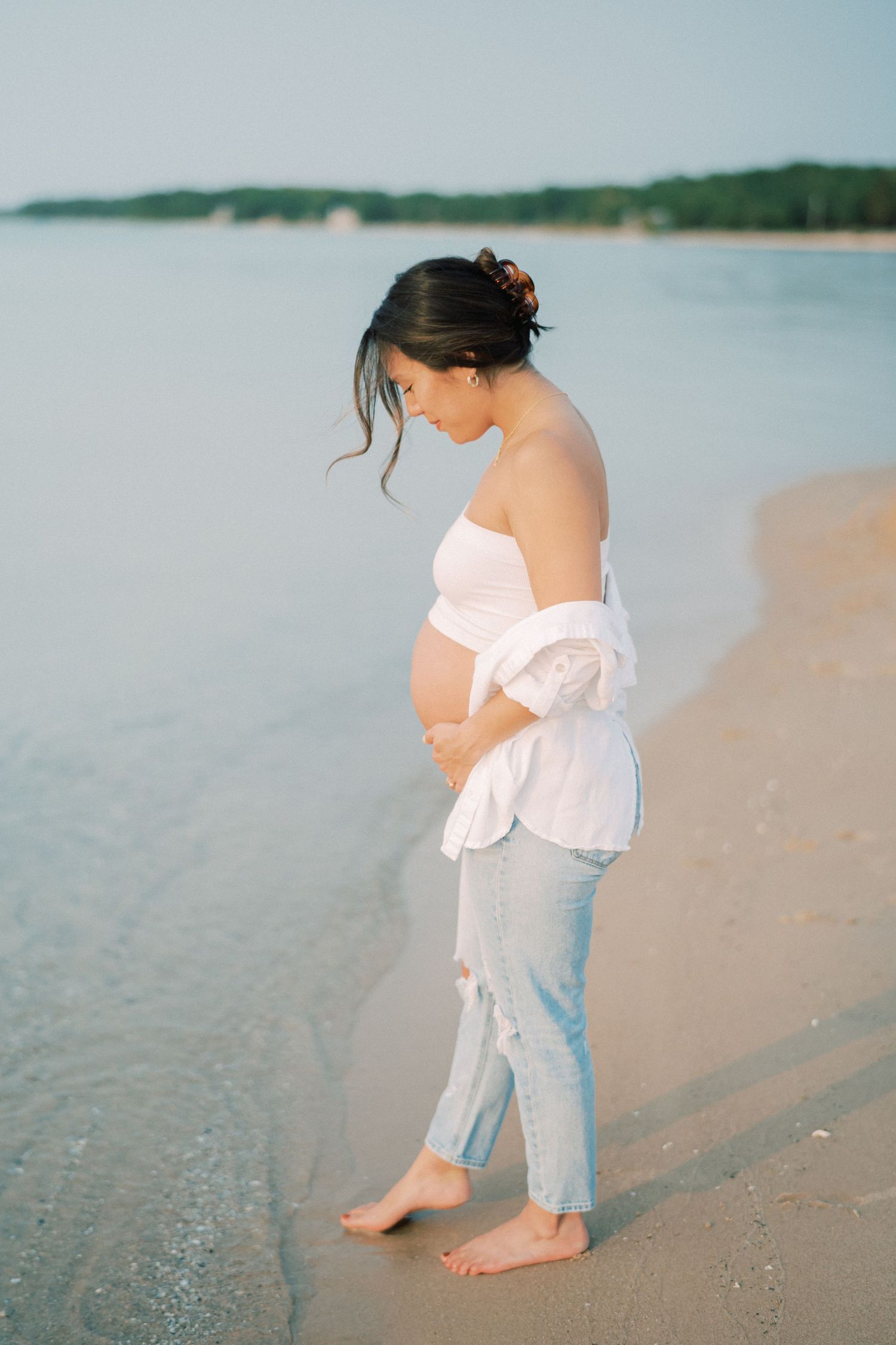 Pregnant woman in white top and jeans walks barefoot along peaceful beach shoreline at sunset.