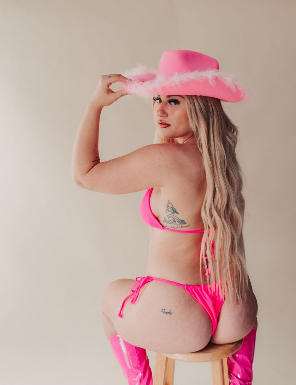 Studio Boudoir Photoshoot
Figure in hot pink cowboy hat and matching bikini poses seated on wooden stool against neutral backdrop.