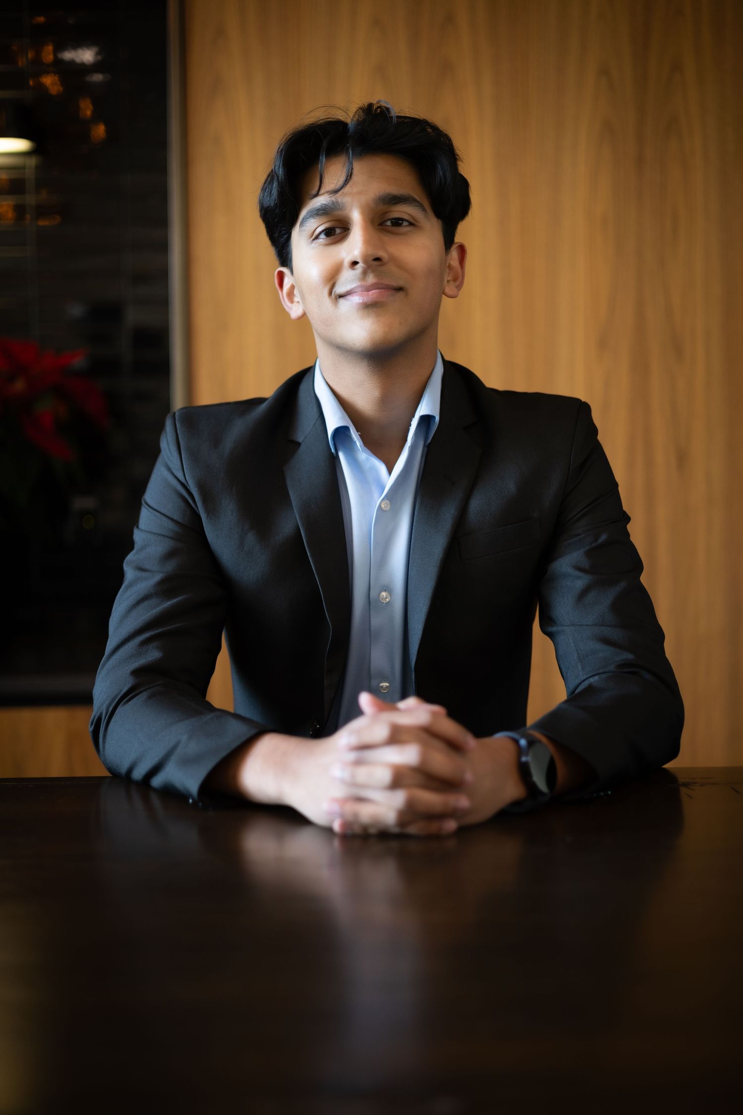Professional portrait of someone in a dark suit and light blue shirt sitting at a wooden table with clasped hands.