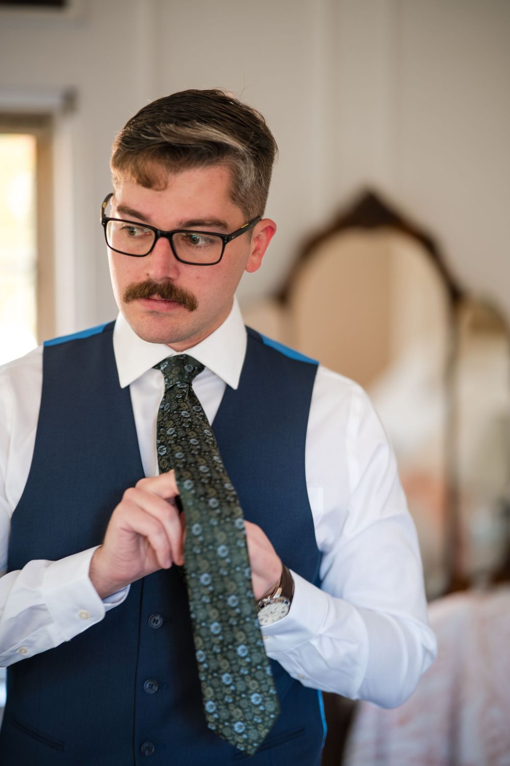 A person in a navy vest and white shirt adjusts their patterned tie while getting dressed.
