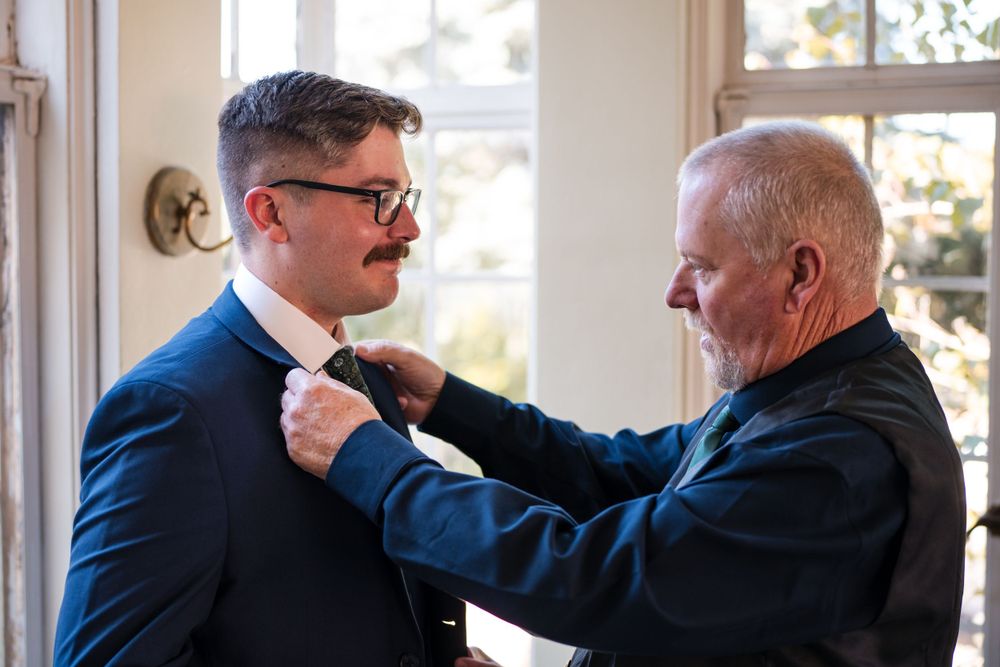 Two people adjusting formal wear near sunlit window.