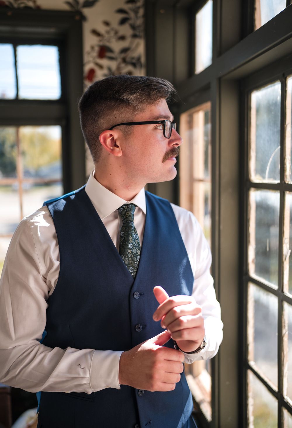 A person in formal attire adjusts their cufflinks while standing near a window.