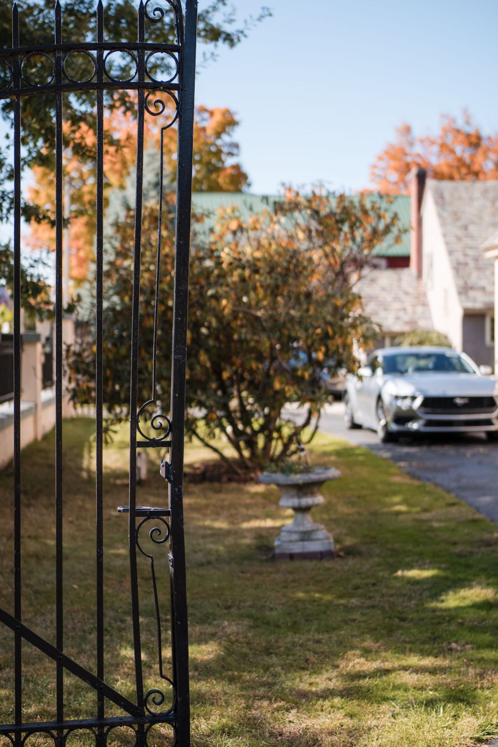 Wrought iron gate opens to reveal a driveway with autumn trees and a parked silver car in the background.