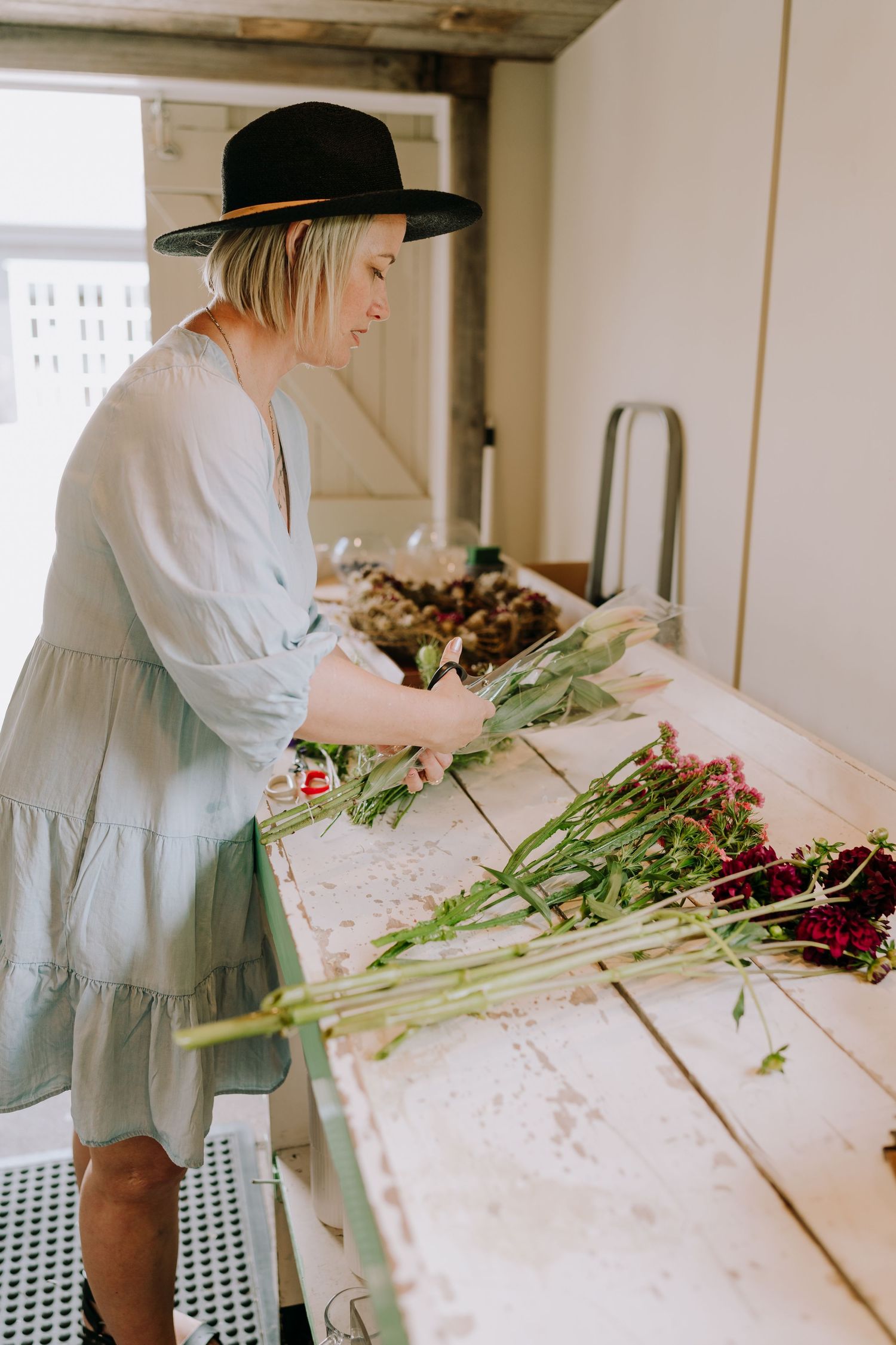 A person in a white dress and black hat arranging fresh flowers on a white table.