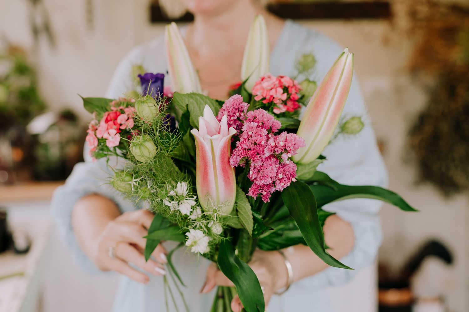 Hands holding a fresh bouquet featuring pink carnations, white lilies, and green foliage.