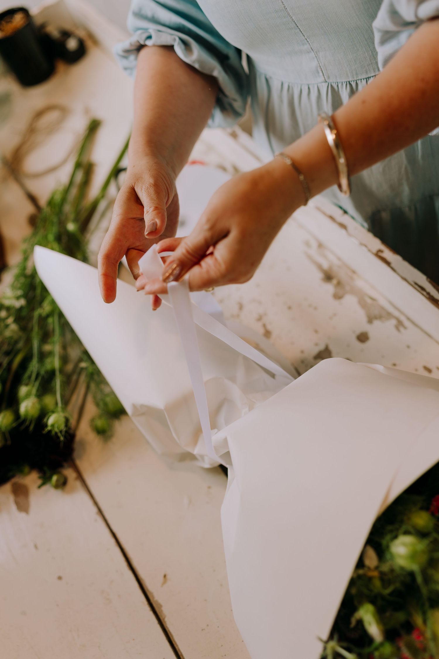 Hands wrapping flowers in white paper on a wooden surface with floristry supplies.