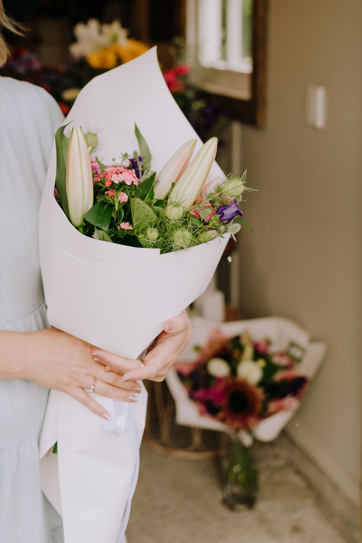 White wrapped bouquet with lilies and pink flowers held against a neutral interior background.