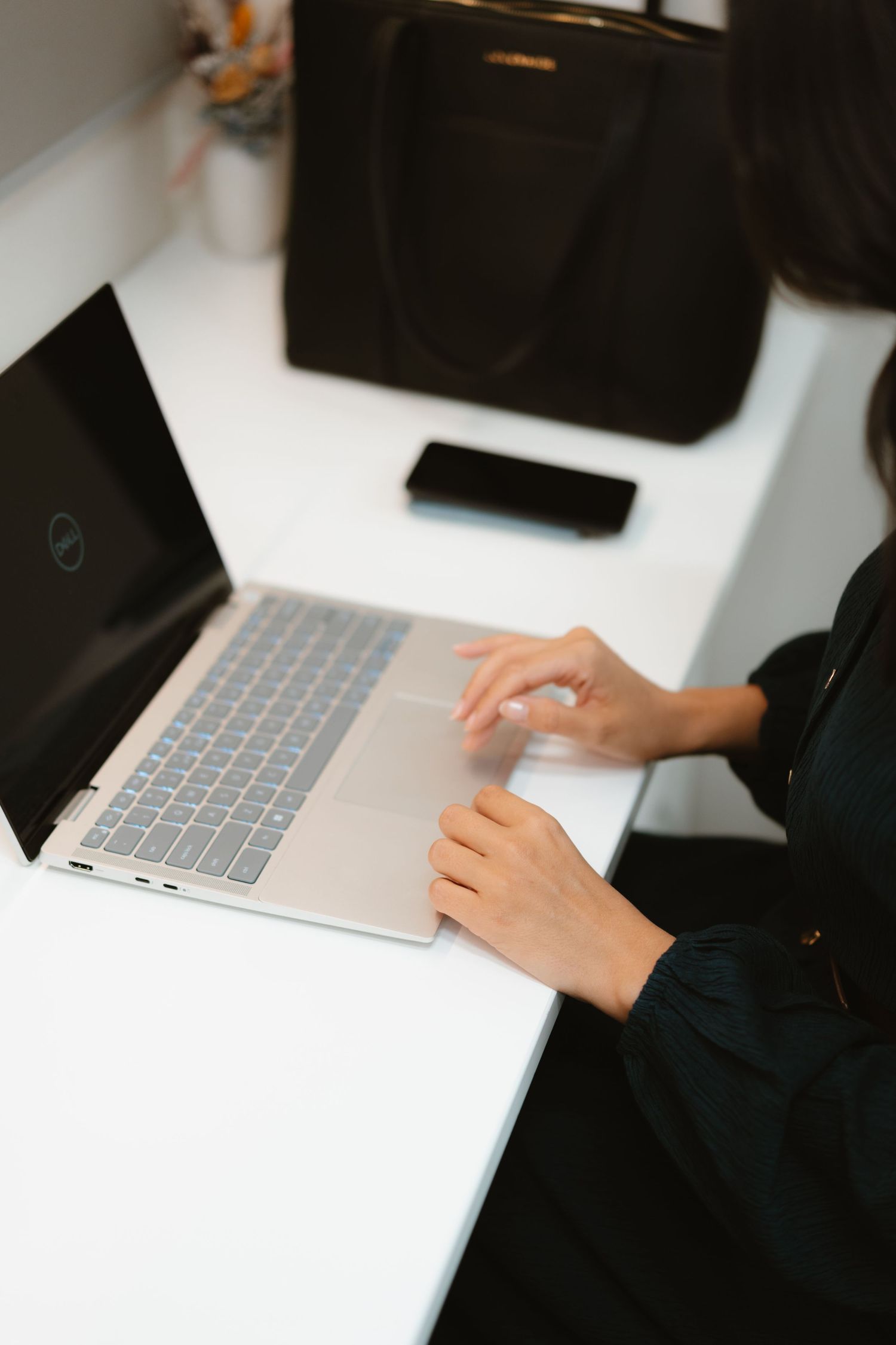 Hands typing on silver laptop keyboard with black bag and phone on white desk.