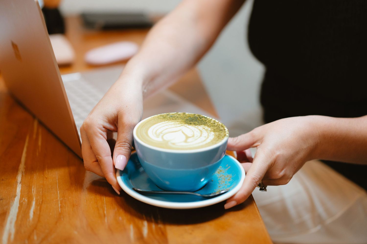Hands serving blue ceramic cup with artistic matcha latte art on wooden table.