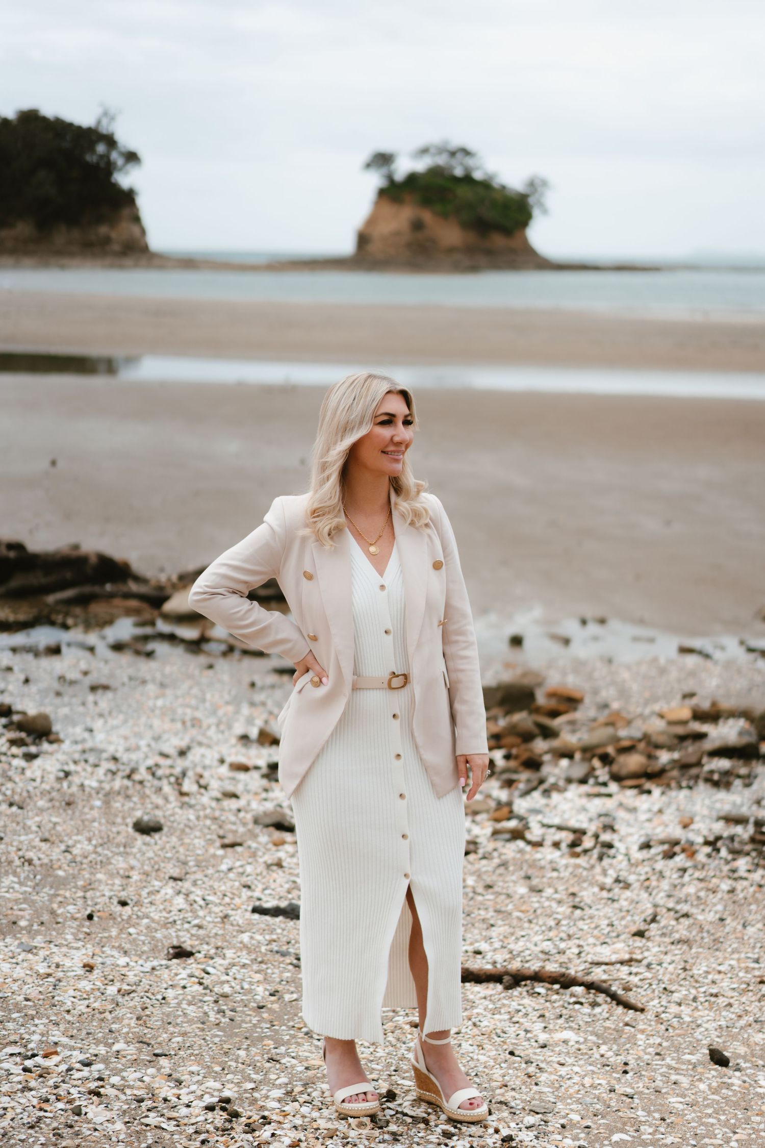 A woman in a white dress and beige blazer poses on a rocky beach with small islands in the background.
