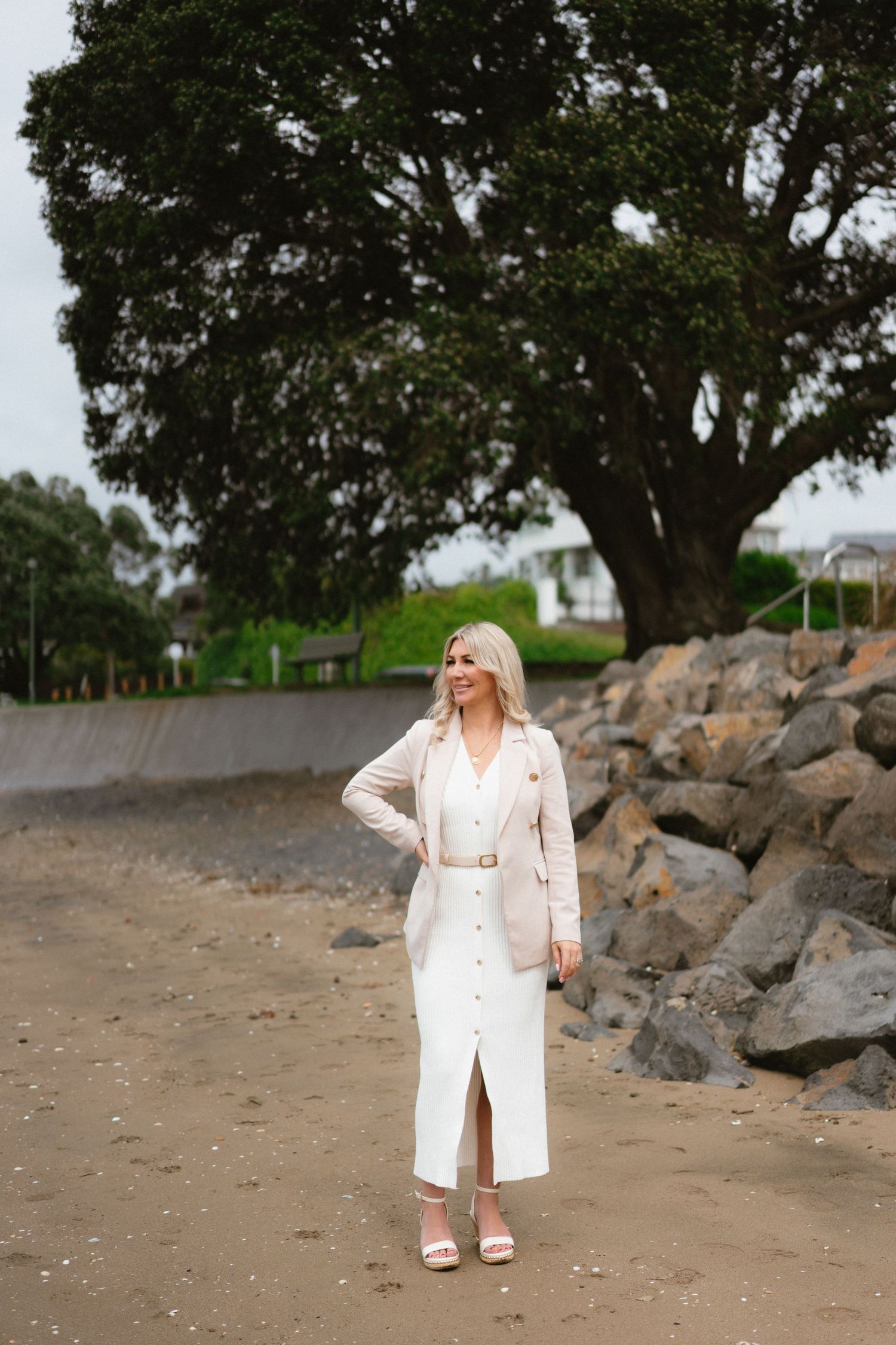 A woman in white outfit stands by large rocks and tree on a coastal path.