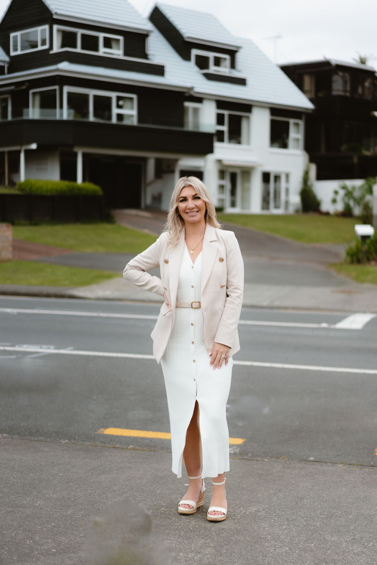 A person in white dress and blazer poses on street in front of a large craftsman style house.
