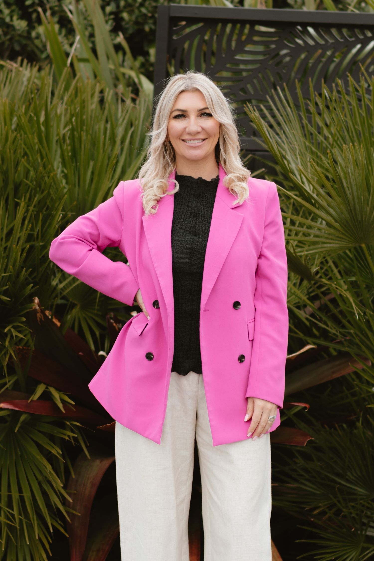 A person wearing bright pink blazer and white pants poses against tropical foliage.