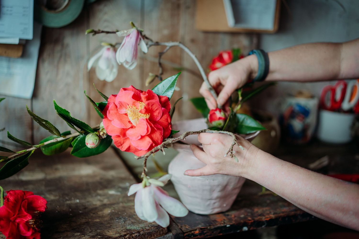 Hands arranging coral flowers and white blooms in rustic setting.