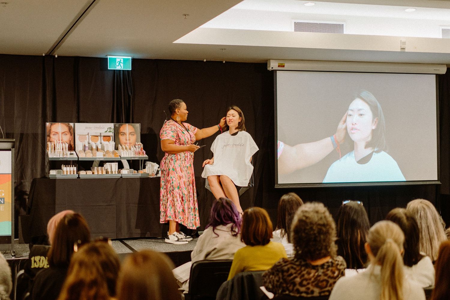 A beauty demonstration taking place at a conference with a projected screen behind the stage.