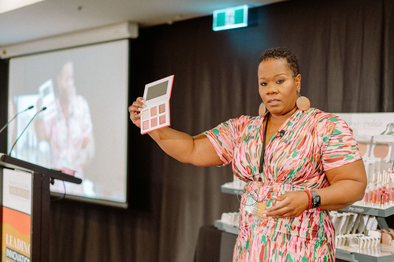 Speaker presenting makeup products at a beauty industry event with presentation screen.