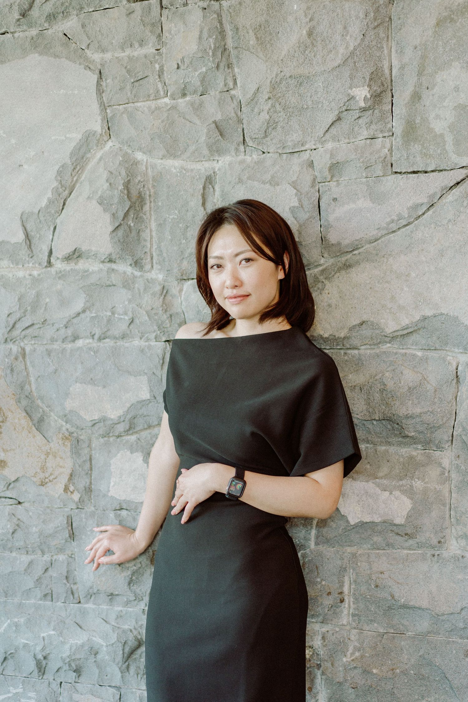 Professional portrait against textured stone wall featuring black dress and natural styling.