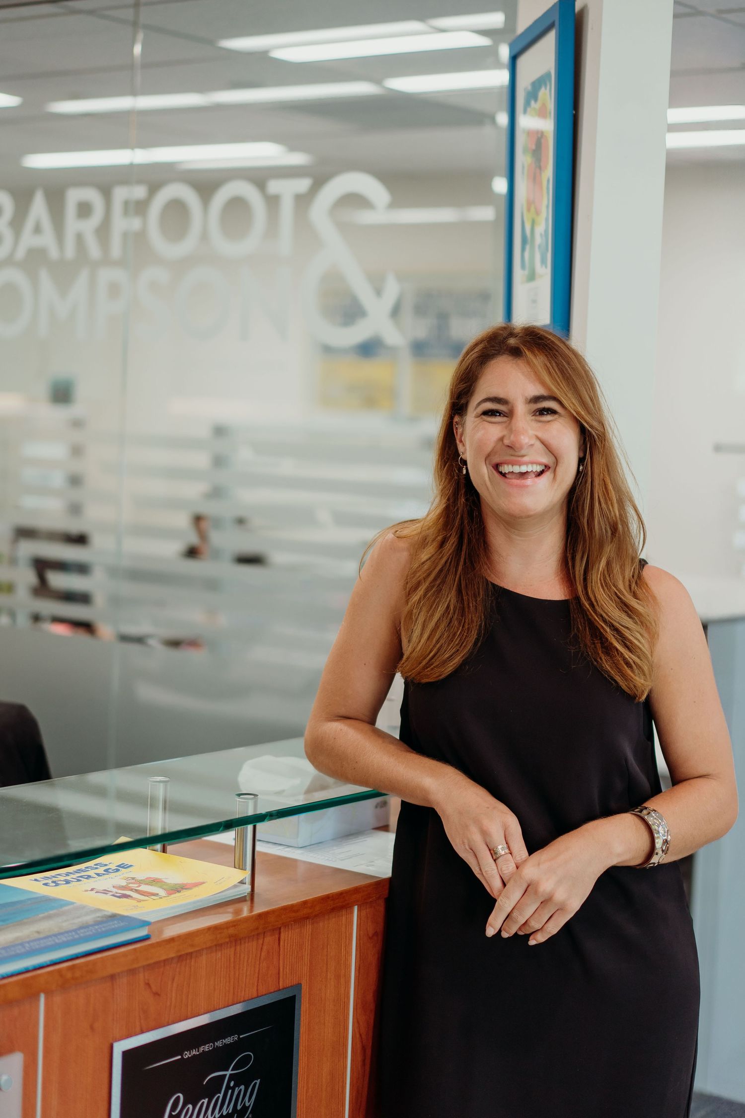 A person in black dress stands at reception desk with Barefoot Thompson signage.