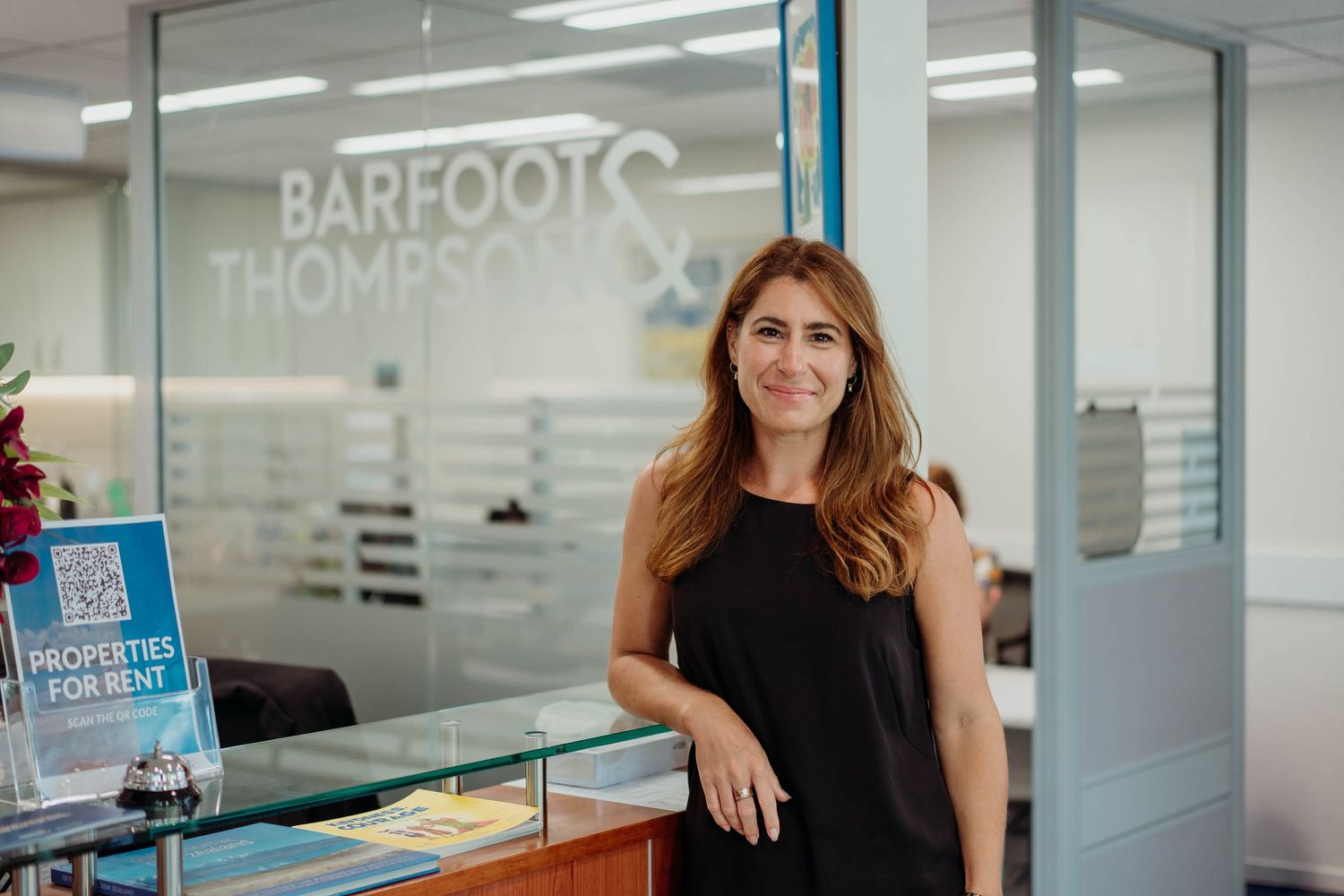 A person in black dress stands in office reception area with glass walls and company branding.