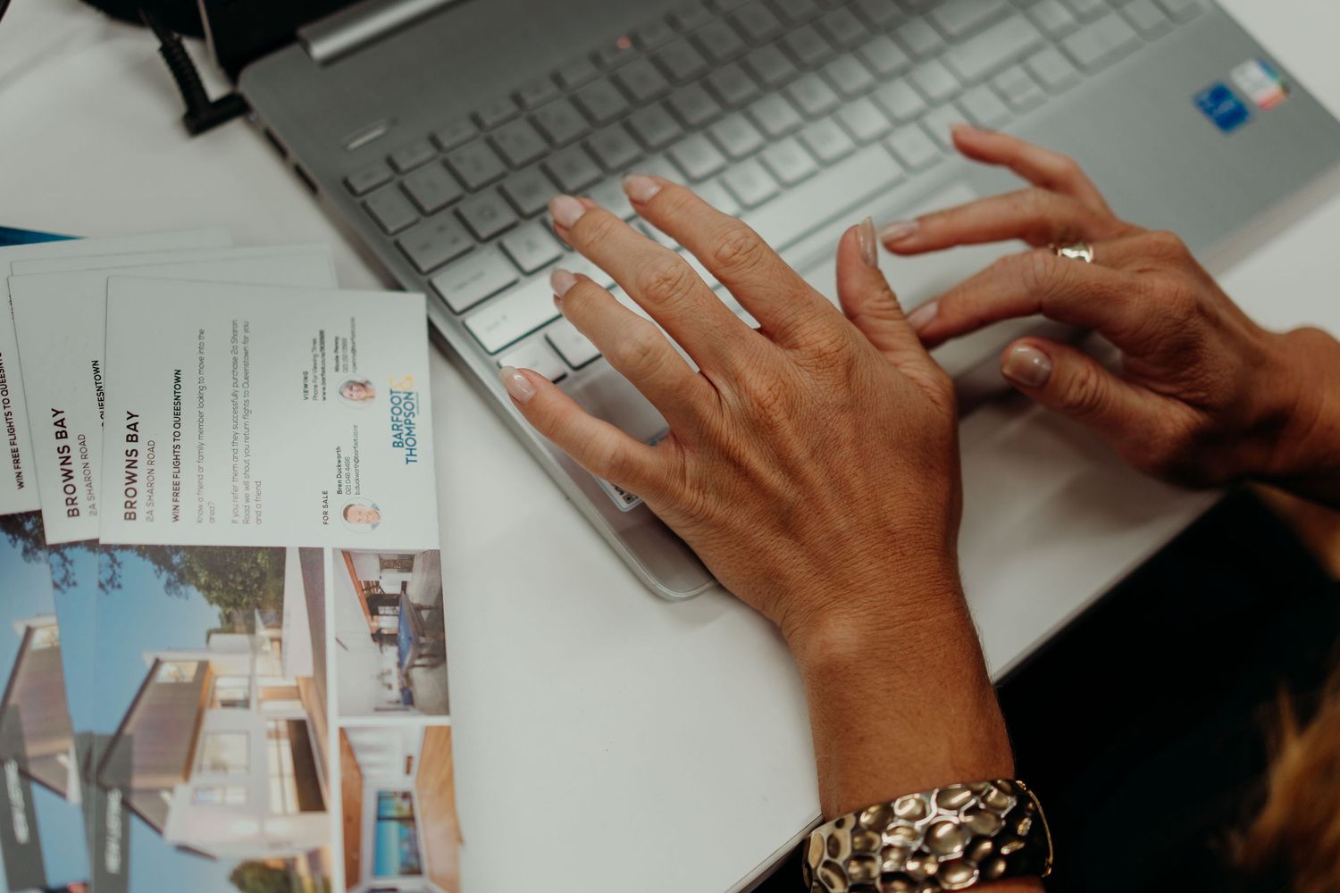 Hands typing on a silver keyboard with jewelry and paperwork visible on the desk.