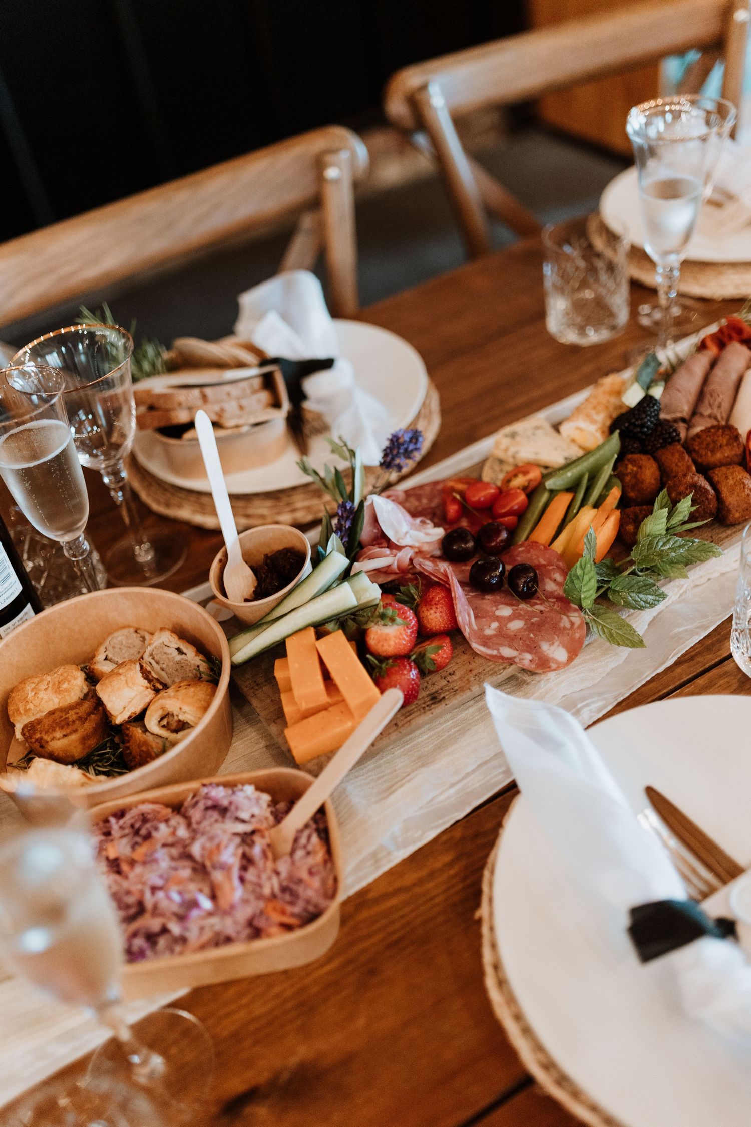 Elevated wooden charcuterie board with assorted meats, cheeses, vegetables and bread served with wine glasses on rustic table.