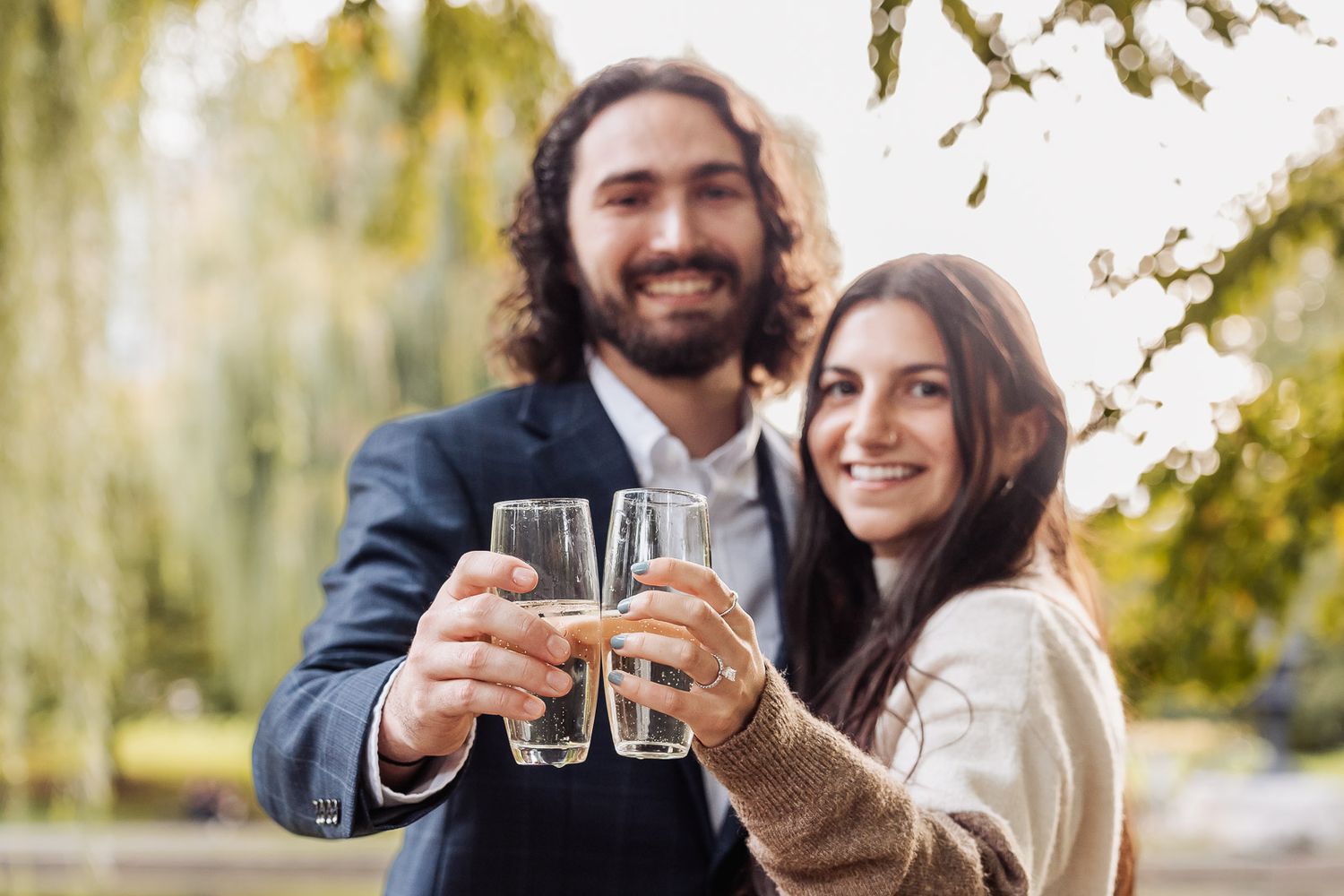 A couple smiling and celebrating with a champagne toast outdoors on an autumn day.