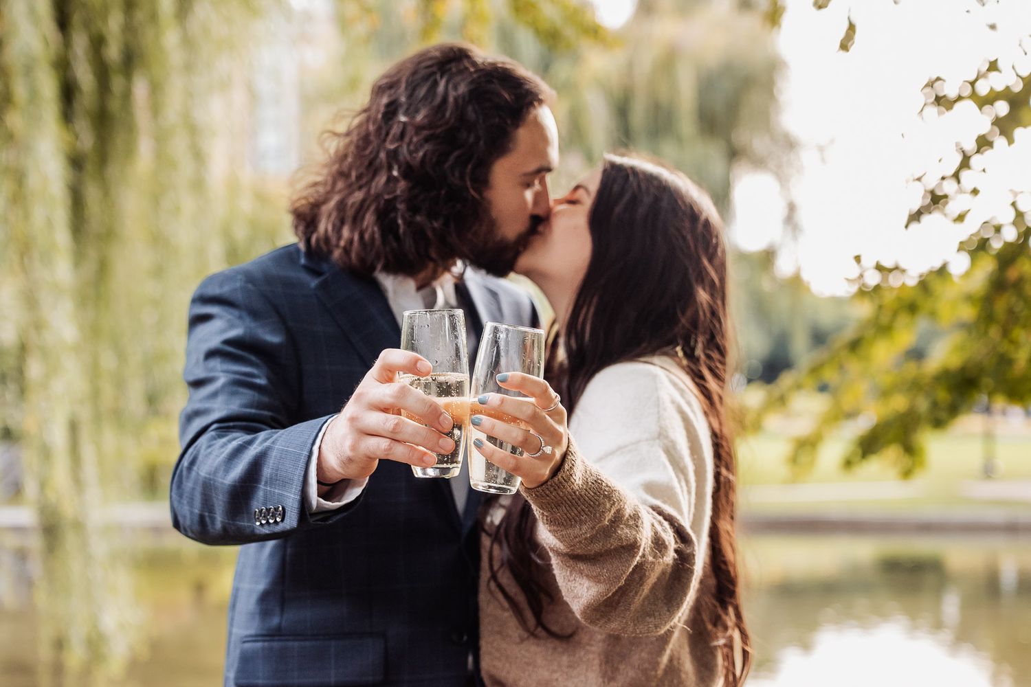 A romantic couple toasting with champagne glasses in a park with willow trees in the background.