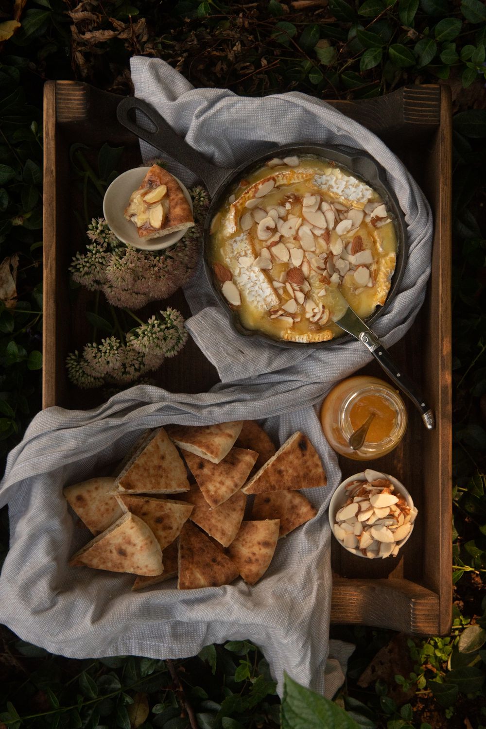 Wooden tray with baked brie, crackers, and almonds served with honey on white linens.
