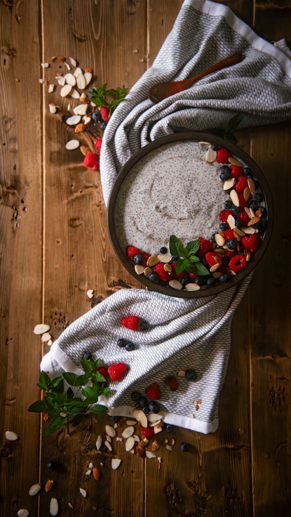 Bowl of smoothie topped with berries, nuts and mint on wooden surface with grey cloth.
