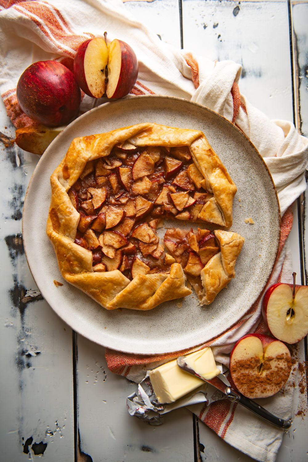 Rustic apple galette with sliced almonds on white plate with fresh apples and butter on wooden surface.