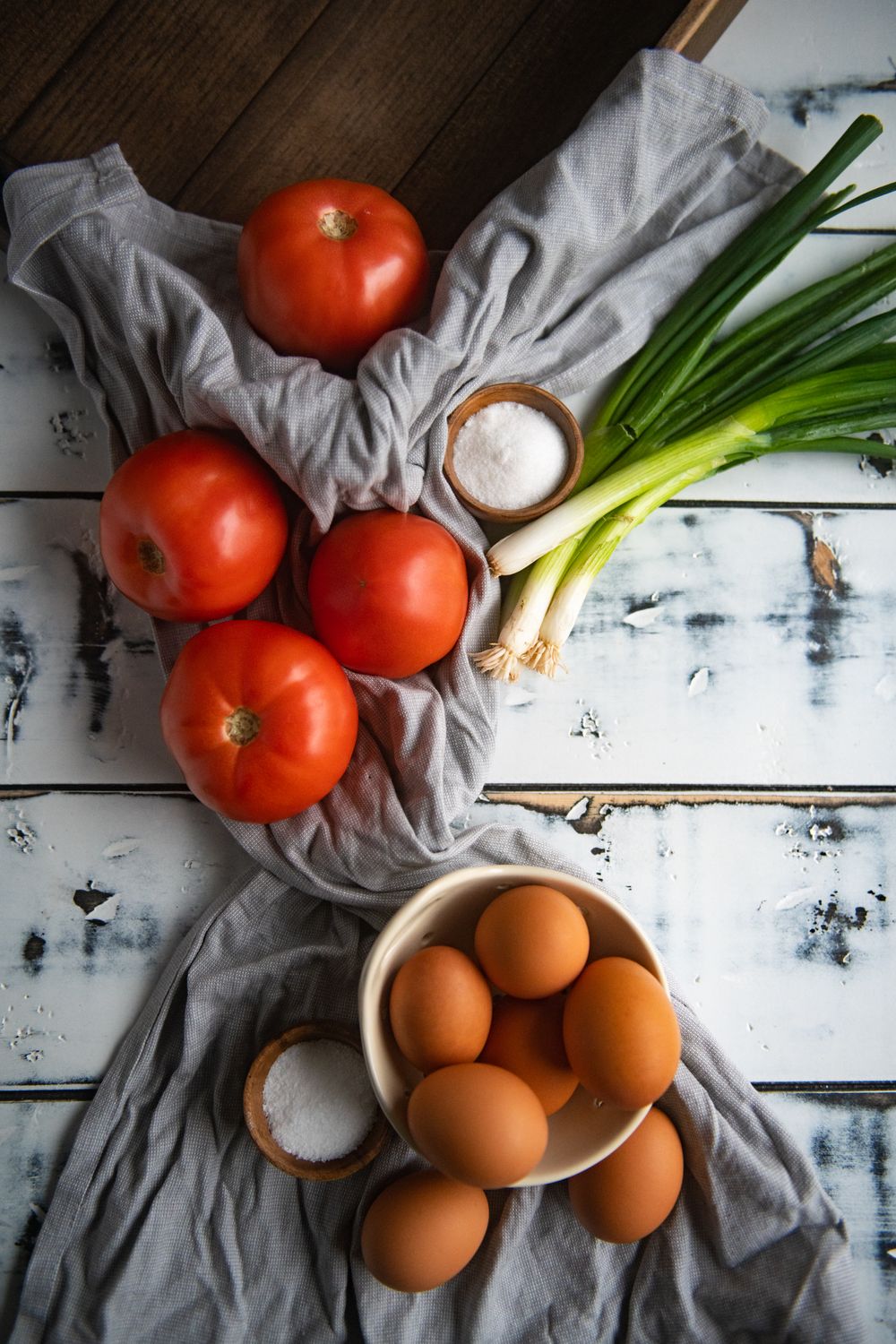Fresh tomatoes, green onions, and brown eggs arranged on grey cloth with white wooden background.