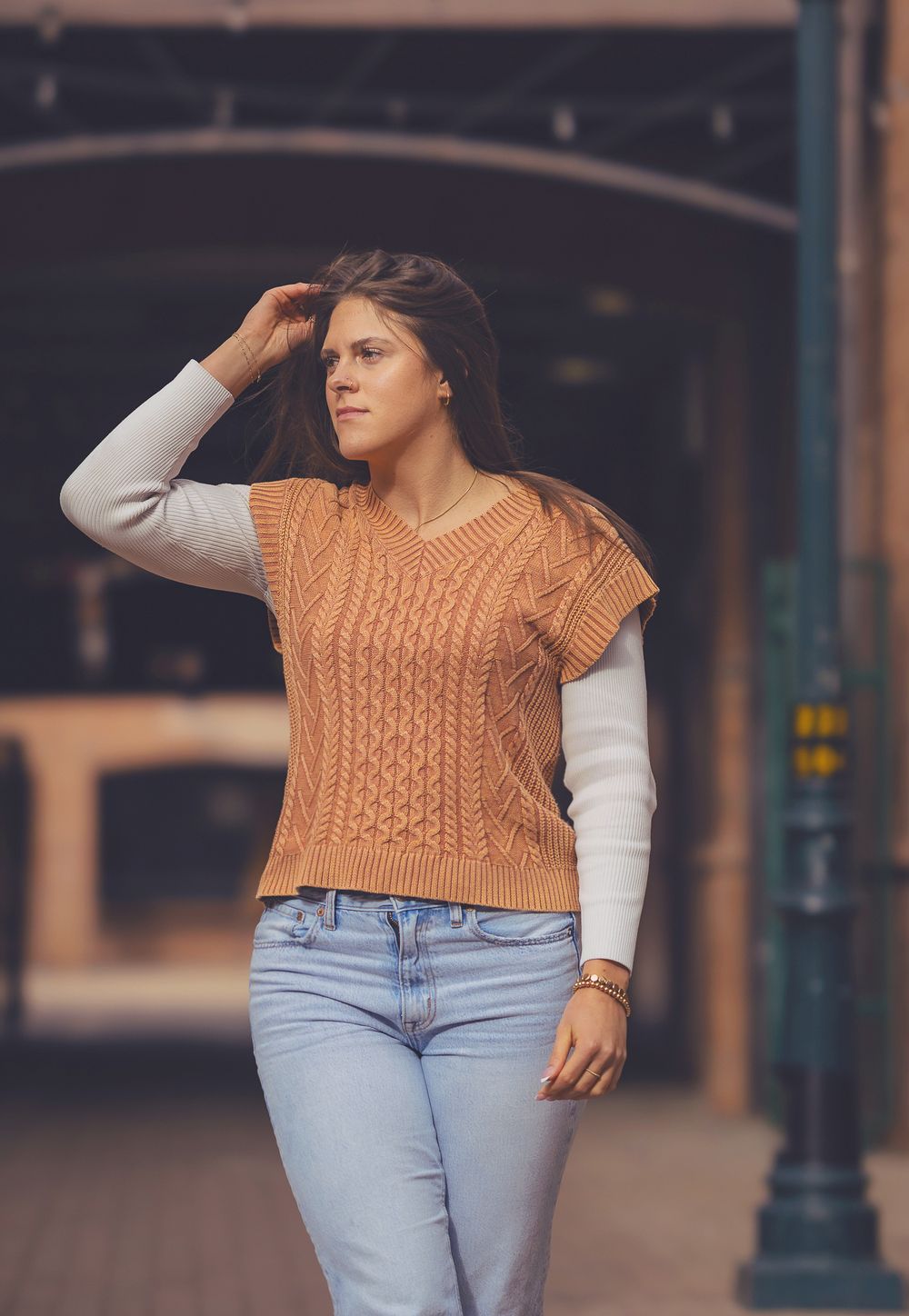 Portrait of a person with long brown hair wearing a peach-colored cable knit sweater against a brick wall background.