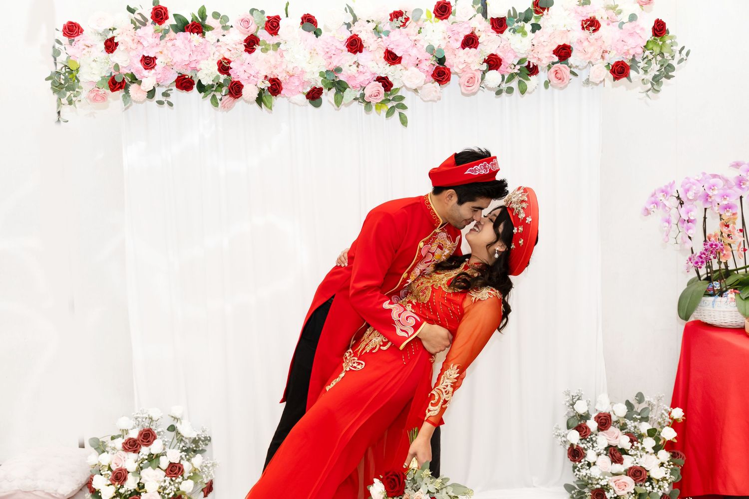 A romantic moment between two people in red traditional attire beneath a floral arch with pink and red roses.