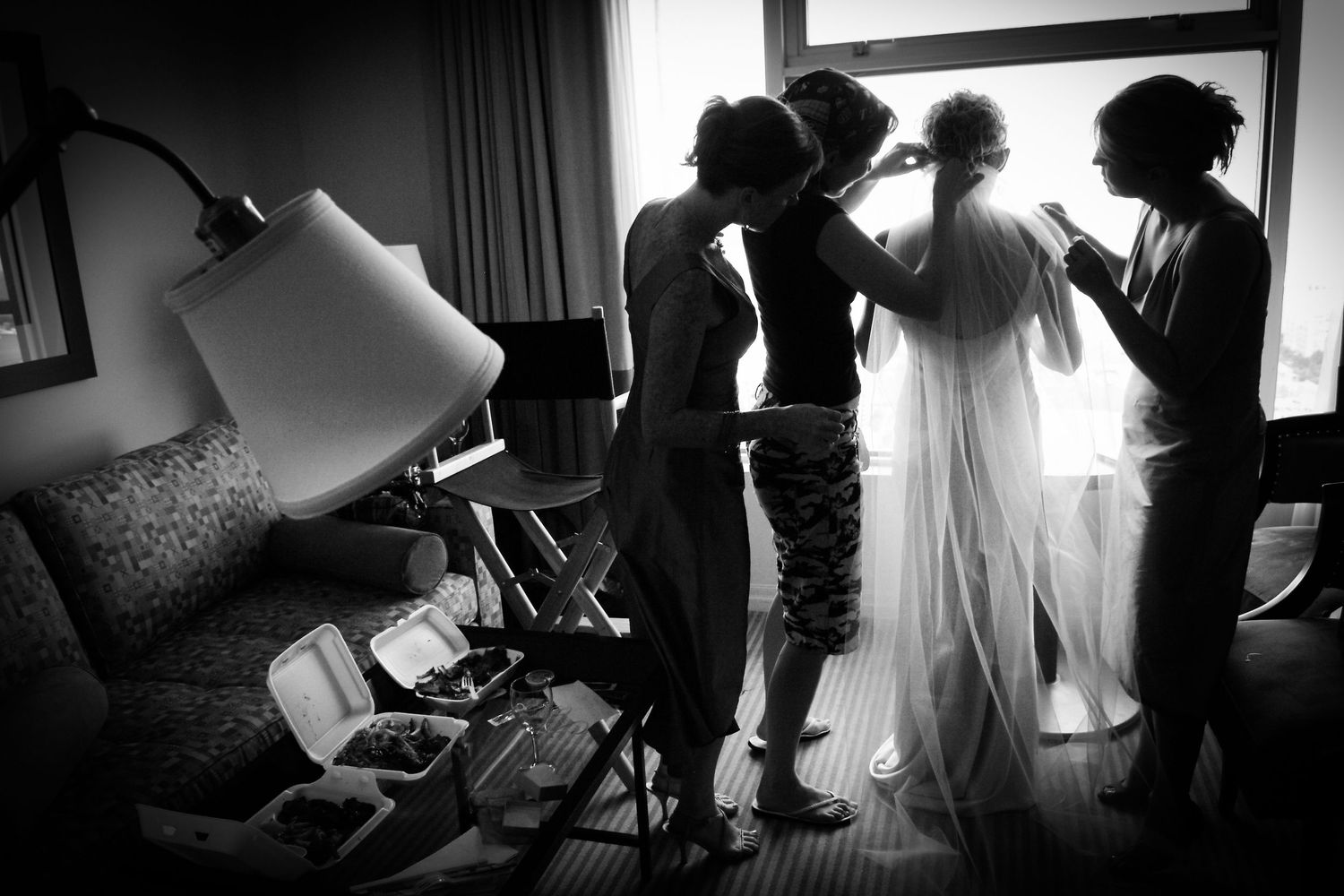 A silhouetted scene of wedding preparation with dress and helpers by a window.