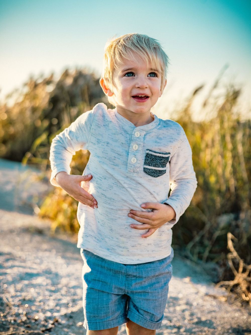 Young child in white henley shirt smiles while holding something on a sunny beach path.