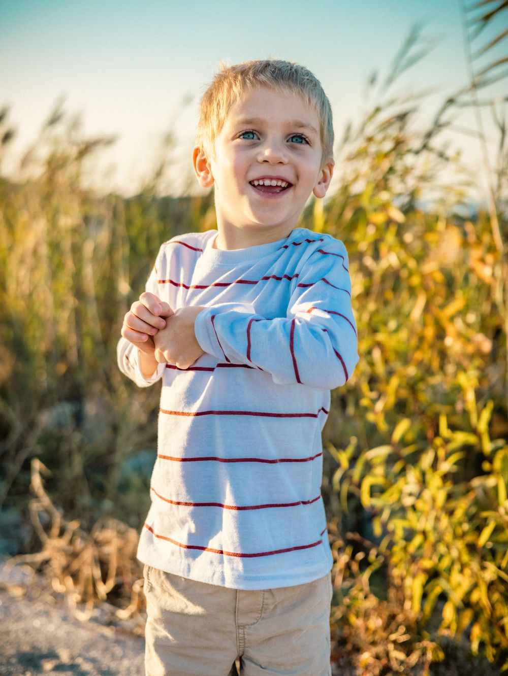 Child wearing striped shirt laughs joyfully in golden sunlight among tall grass.