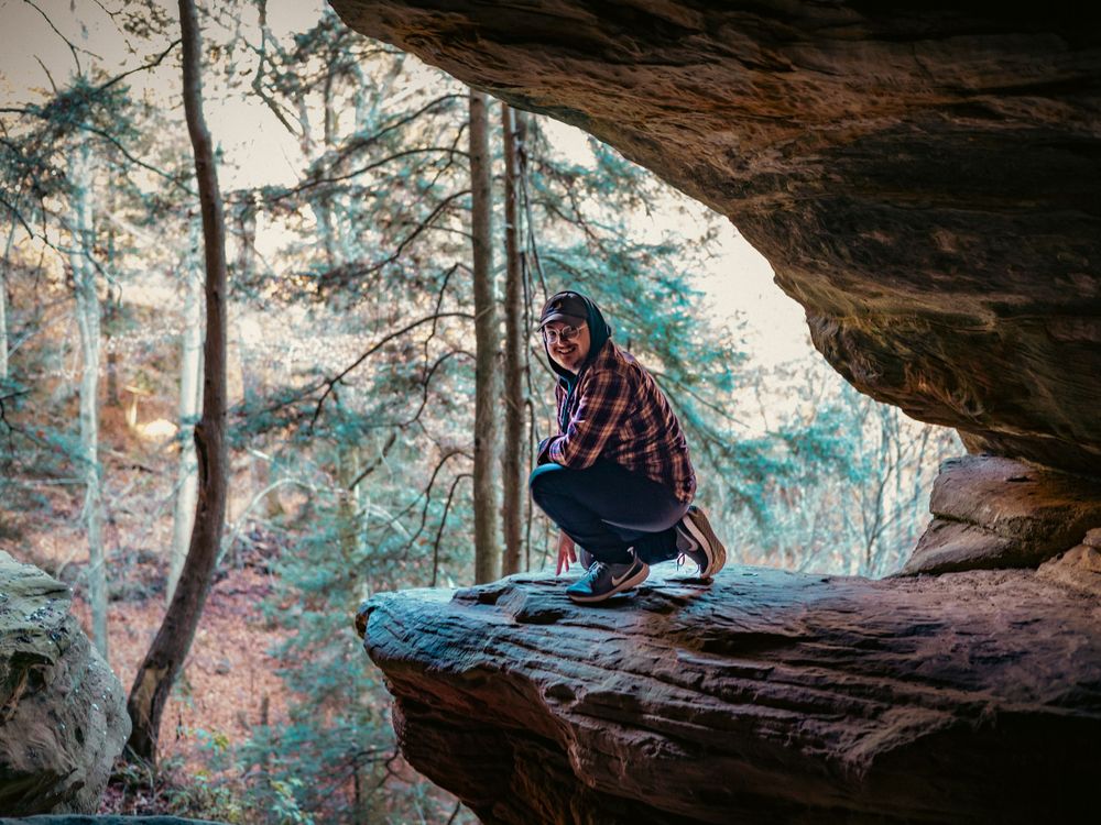 Adventurer crouching at edge of cave opening overlooking forest landscape.