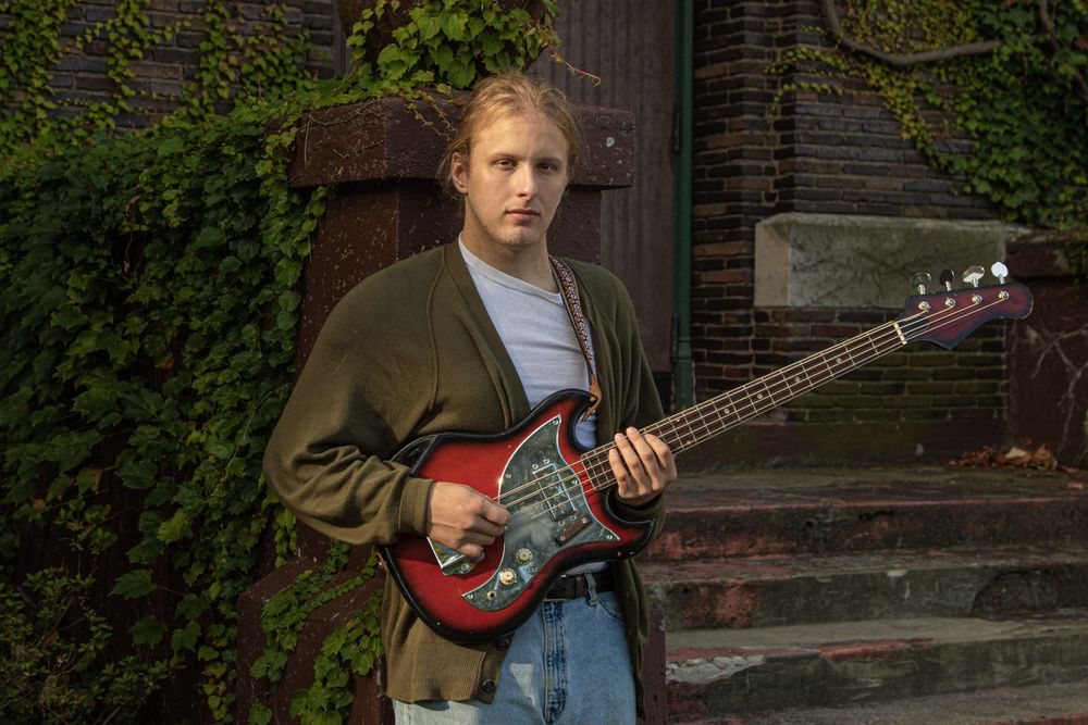 Musician holding red electric guitar standing near ivy-covered wall.