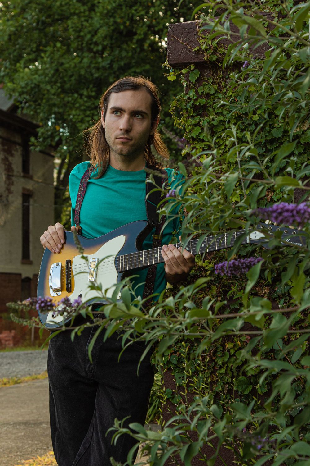 Musician with long hair playing white electric guitar outdoors near garden.