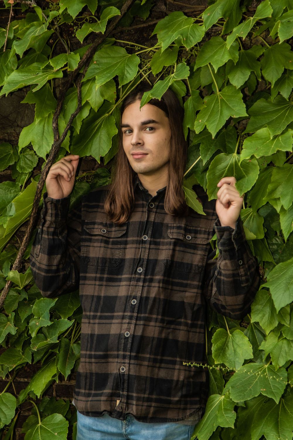 Person in plaid flannel shirt poses against green ivy-covered wall in outdoor portrait.