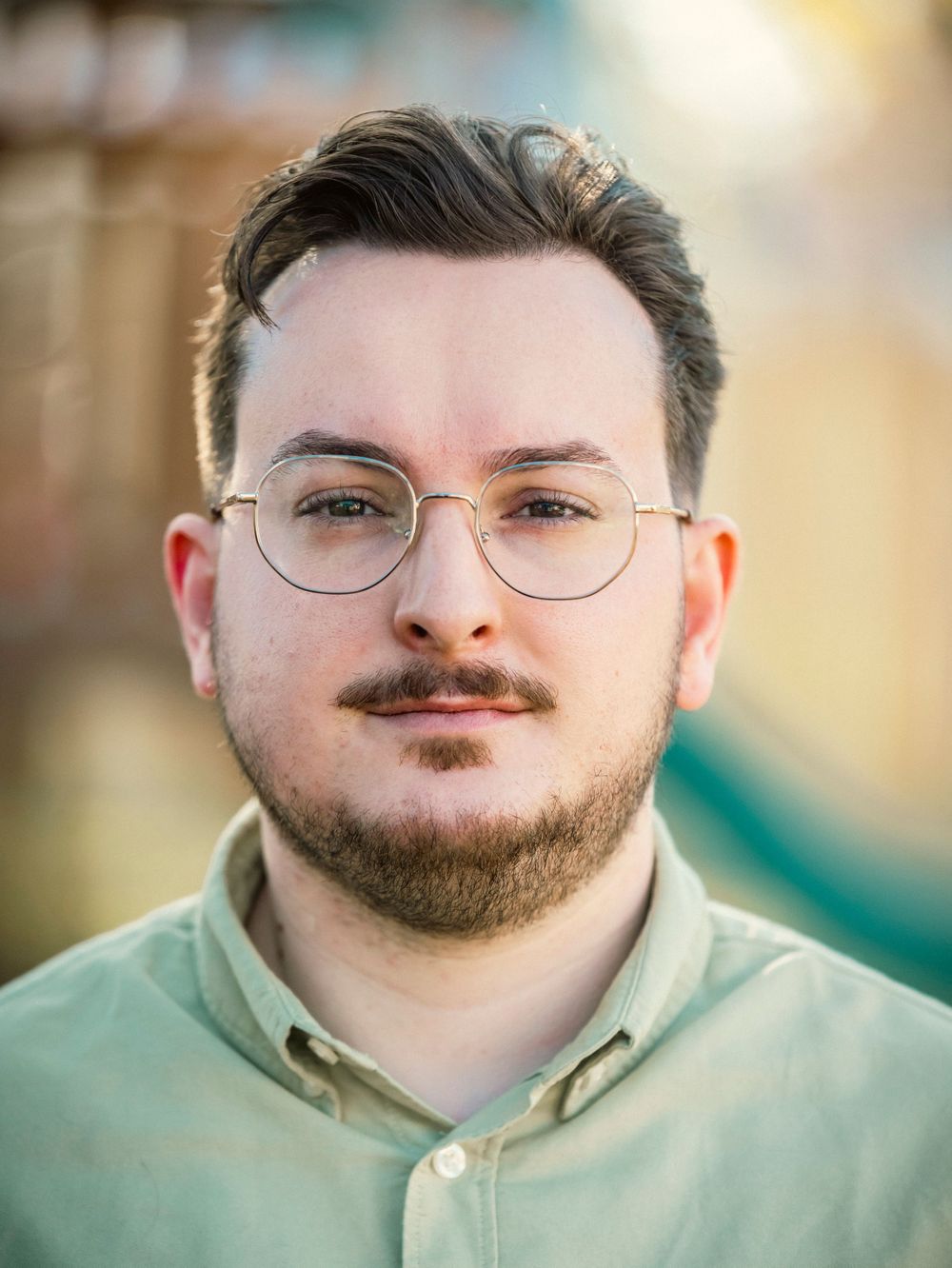 Portrait against blurred backdrop wearing glasses, light shirt and neat facial hair.