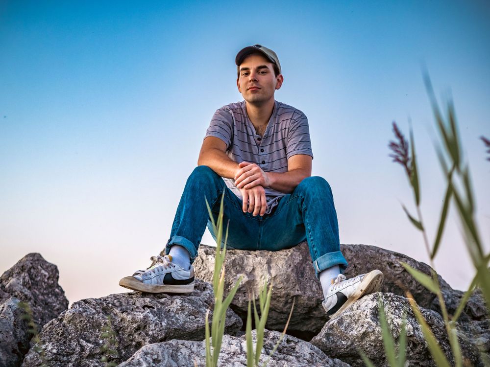 Subject sitting on rocky outcrop with grass and blue sky in background.