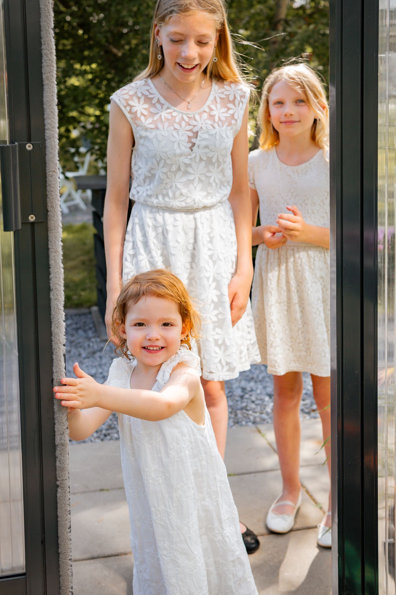 Tre tjejer i vita spetsklänningar står i dörren till trädgårdens växthus en solig sommardag. Familjefotograf, Umeå, Fotograf, Västerbotten