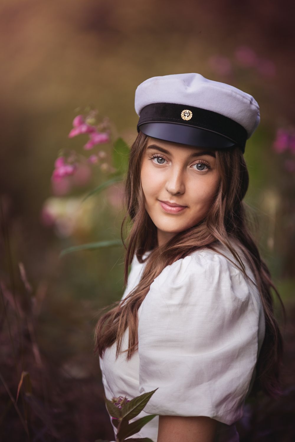 Portrait of someone wearing a white nautical cap and white blouse among blooming pink flowers in soft outdoor lighting.
