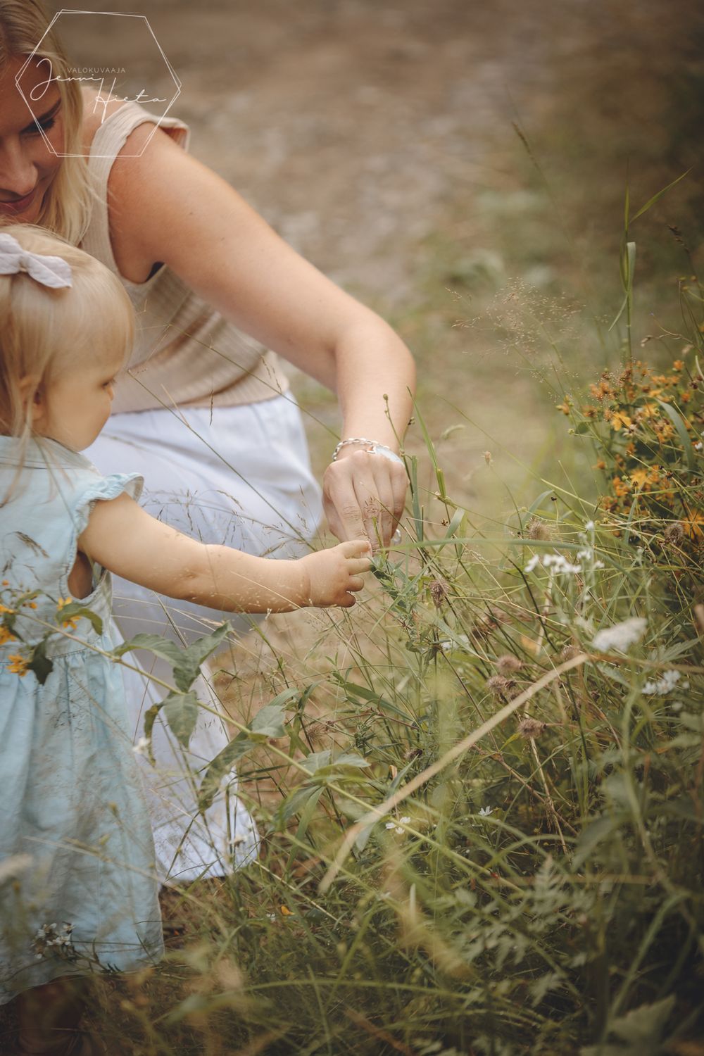 A gentle moment in nature as a young child in a blue dress explores wildflowers with guidance in a dreamy outdoor setting.